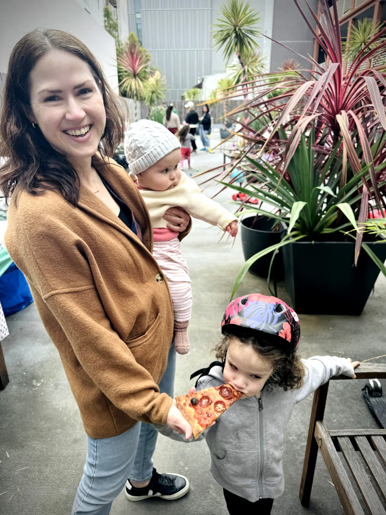 Constance holding her baby daughter while her older daughter, in a bike helmet, eats a slice of pizza